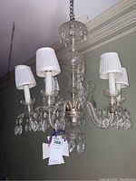 Full view of the glass chandelier from below, showing six white candle-style light bulbs with pleated white fabric shades and hanging crystal prisms.