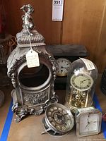 Group view of four clocks: silver mantel case, black wooden mantel clock, Linden anniversary clock, Seiko Quartz tabletop clock