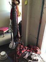 Photo showing brass coat tree with multiple hooks, hanging a black and white patterned scarf and other garments. Red and black plaid jacket and other clothes on the floor.