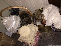 Porcelain doll in a pink dress with bonnet and ribbons, doll wig, sunhat, lace lampshade, woven basket, brass pot, and purses arranged together