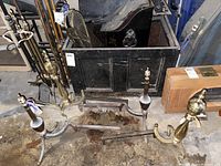 Photo showing one pair of brass and iron andirons with curved bases and finials, black metal ash box, and part of a fireplace fence.