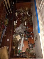 Drawer filled with brass colored lamps, silver plate candle holders, glass door knobs, and electrical cords inside a wooden drawer.