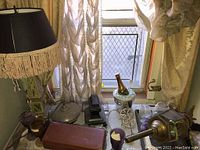 Photo of lamp, brass pot, glassware, cheese boards, serving trays, and other items on a table by window