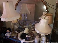 Four table lamps on a table with scalloped cream shades and decorative bases, a blonde doll in blue dress, lamp shades and candle sticks visible