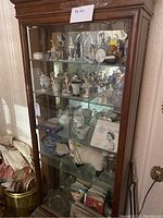 Full view of wooden display cabinet with glass shelves showing porcelain figurines, teacups, plates, a stuffed sheep, books, and vases.