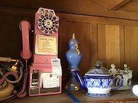 Photo showing pink rotary dial payphone, blue and gold urn, blue and white teapot with lid, and white figurines on wood background.