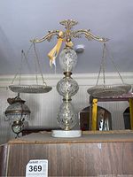 Front view of the full balancing scale showing the glass central post, brass balance beam with ornate grape motifs, and two glass weighing pans suspended by chains.