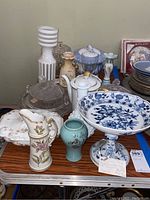 Overview of glass and porcelain serving items on a table featuring platters, pitchers, vases, bowls, and candlestick holders in various styles and colors.