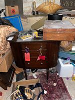 Vintage sewing table with a drawer, dark wood, tapered legs, and curved front panel with two wooden knobs. Located in a basement area with some clutter around.