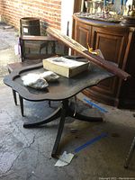 Wooden table with scalloped edges, a wooden plant stand, slippers, and mahogany wall shelf visible in background, all displayed in garage with concrete floor and brick wall.