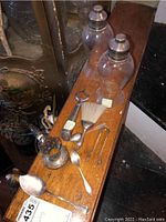 Photo showing two cocktail shakers with sterling silver tops, a brush with silver handle, small tongs, bottle opener with faceted handle, spoons, and etched glass piece holders arranged on a wooden surface.