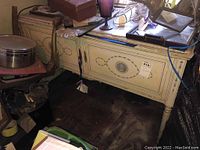 White painted wood table with marble top and decorative painted cabinet base shown from a side angle with other household items surrounding it.
