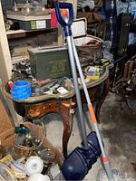 Photo of snow shovel, mop, and green military metal ammo box on an antique curved wooden table in basement with surrounding items.