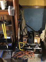Photo showing shovels with yellow handles, extension cord, paint cans, hand tools, and a toolbox in a garage corner