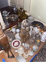 Various vintage and decorative items including glassware, ceramic tea cups, silver pitchers, and brass candle holders arranged on a wooden surface.