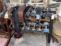 Dumbbells ranging from 5 lb to 30 lb arranged on a black metal dumbbell stand with several leather weight lifting belts draped on top. The stand is located in a basement with a stone wall background.
