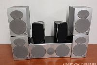 Five Athena surround sound speakers arranged with two smaller bookshelf speakers in the center on a wooden surface against a white background.