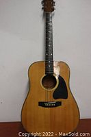 Full front view of acoustic guitar showing natural wood finish, black pickguard, six steel strings, and a wooden bridge.