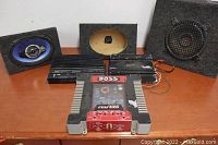 Wide shot of all car audio equipment on table showing 3 amplifiers, 2 speakers in carpeted enclosures, and one empty speaker enclosure box.