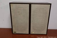 Pair of Genesis floor speakers shown front-facing with fabric grills and dark wood frames on a wooden surface against a white wall.