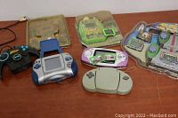 Photo showing six handheld and TV plug-and-play game devices of various shapes and colors arranged on a wooden surface.