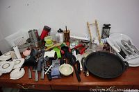 Photo showing many kitchen accessories laid out on a table including graters, peelers, knives, white plastic storage containers, spatulas, a large black metal griddle pan, and assorted utensils.