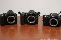 Three Nikon DSLR cameras D5000, D80, D40 side by side, without lenses, on wood table.