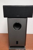 Front view of the subwoofer and center channel speaker stacked, showing black fabric grille on speaker and circular port on subwoofer with control panel below.