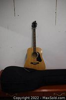 Full view of Rocker acoustic guitar standing against wall with soft case lying in front.