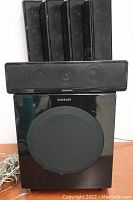 Photo showing four slim black Samsung satellite speakers vertically aligned behind a black Samsung center speaker and a black subwoofer below with circular front speaker grill.