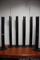 Six black tower speakers standing upright, four identical Panasonic branded and two Samsung speakers on wood surface near white wall.