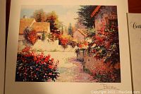 Close-up view of the art print showing a village scene with flowers and houses, artist's signature in lower right corner