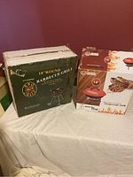 Photo showing two boxed portable grills: an 18-inch round barbecue grill with wheels and a 14-inch portable charcoal case grill.
