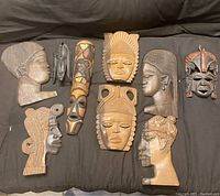Nine African wood art pieces arranged on black fabric including masks and carved heads, showing front views and styling details.