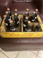 Image showing twelve vintage Coca-Cola glass bottles with metal caps arranged in a yellow wooden Coca-Cola carrying crate with red branding and holding compartments.