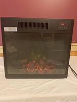 Front view of electric heater showing glass front with illuminated fire log effect and control panel on top right, placed on a flat surface against red wall.