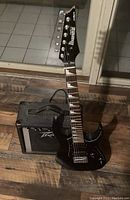 View of the black Ibanez electric guitar standing against the Peavey amplifier on wooden flooring with a tiled floor background.