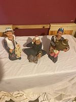 Three handmade figurines of elderly people in traditional attire, displayed on a white cloth against a wooden background.