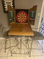 Metal frame rectangular high top table with natural wood plank top and dartboard set on tabletop, two metal chairs with cushioned seats beside it