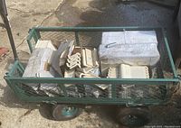 Photo of green metal utility cart loaded with tile boxes and L-shaped tile edge pieces inside on wheels outdoors on pavement.