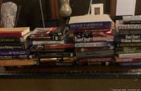 Stack of approximately 20-30 hardback books arranged horizontally on a shelf, showing a variety of titles.