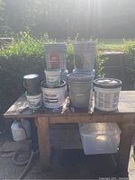 Multiple paint buckets and Sheetrock mud containers in various sizes and colors on wooden table outside, good used condition.