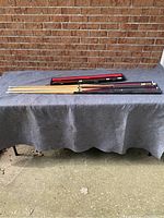 Lot of seven pool cues laid horizontally on a table covered with gray fabric against a brick wall