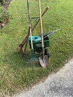 Photo shows multiple yard tools including a green Scotts seed spreader, shovel, rake, and other wooden handled metal garden tools arranged on grass near pavement.
