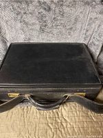 Photo of the closed black leather briefcase showing the top and handle with brass hardware.