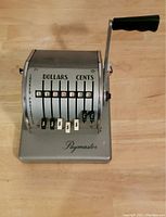 Top view of vintage Paymaster manual check writing machine on wooden surface, showing dollar and cent value tabs and crank handle.