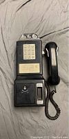 Vintage black payphone with numeric push-button dial, coin slot, and handset with coiled cord placed on fabric surface.