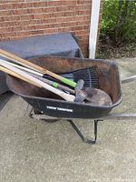 Rusty True Temper wheelbarrow containing yard tools with wooden handles and metal heads inside, placed on concrete near a brick wall