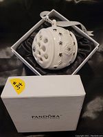 Side view of the white porcelain ornament showing star cut-out patterns and silver accents, hanging ribbon inside Pandora box.