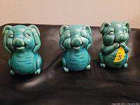 Front view of three turquoise ceramic pig figurines representing 'See no evil, Hear no evil, Speak no evil'. Each pig displays distinct hand placement on facial features.
